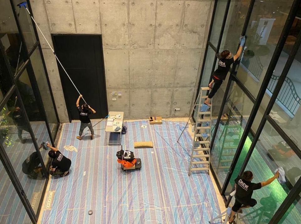 OCD crew cleaning floor-to-ceiling glass panels in a double-height space, with the floor covered in protective sheeting