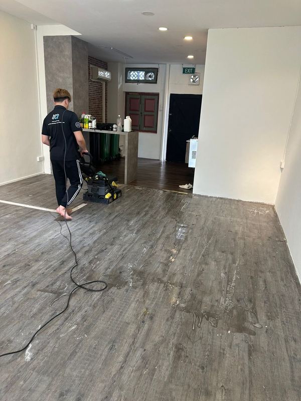 OCD crew member running a walk-behind floor scrubber across a grey wood-look vinyl floor in an empty commercial unit