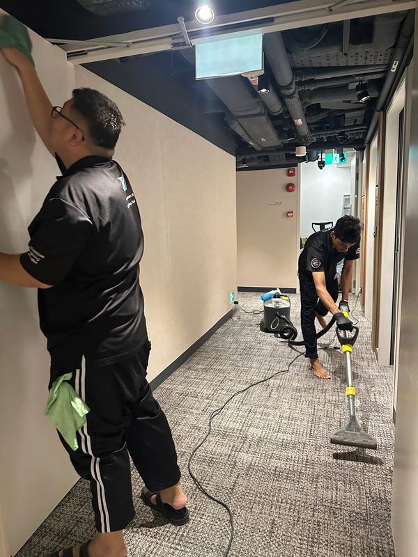 Two OCD crew members in a carpeted office corridor, one wiping down the wall panels and the other running a carpet extraction wand